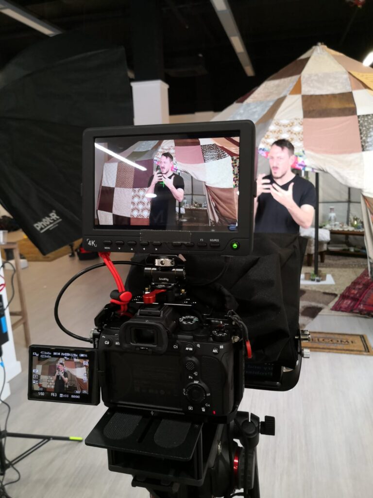 Camera with a telprompter being used. A person is standing in front of the camera.